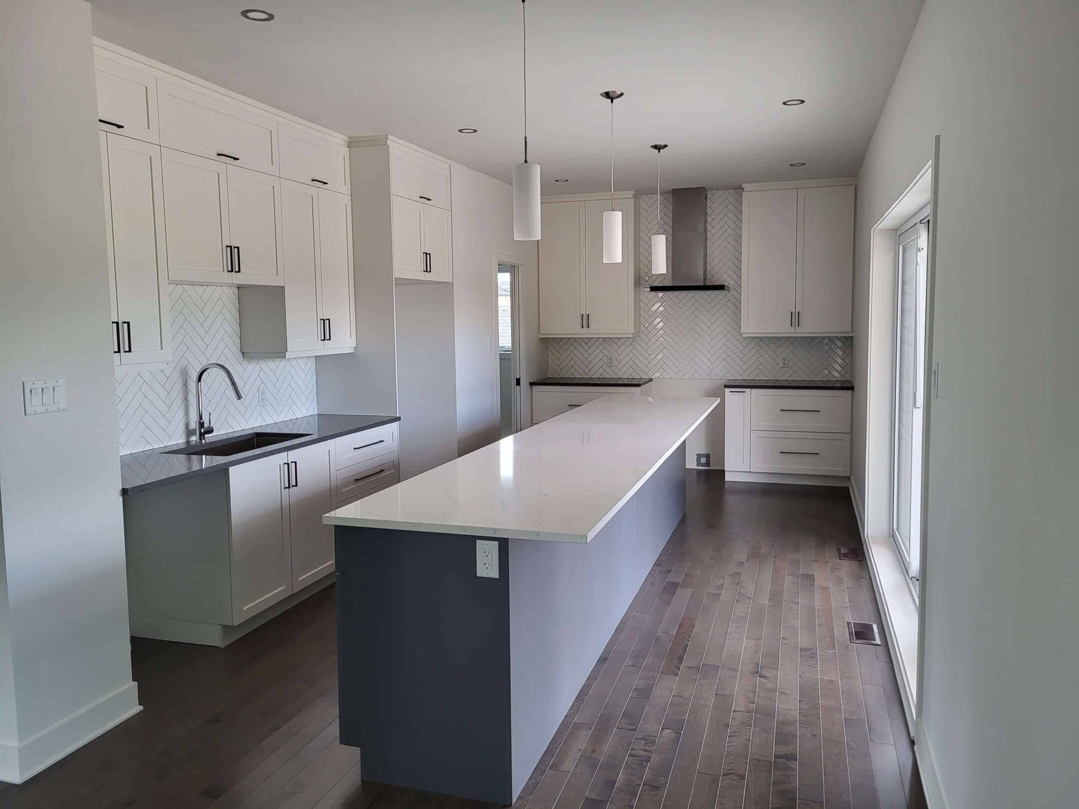 maison neuve pointe aux trembles Cuisine moderne d’une maison neuve au Québec avec îlot central, armoires blanches et dosseret en chevrons, exemple de finition haut de gamme d’un constructeur résidentiel.